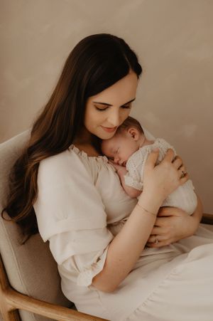 Tender moment between mother and sleeping baby in white outfits against neutral backdrop.