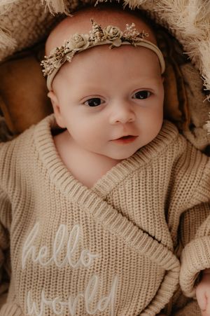 Baby wearing a knitted cream sweater and delicate floral headband looking at camera in warm toned portrait.