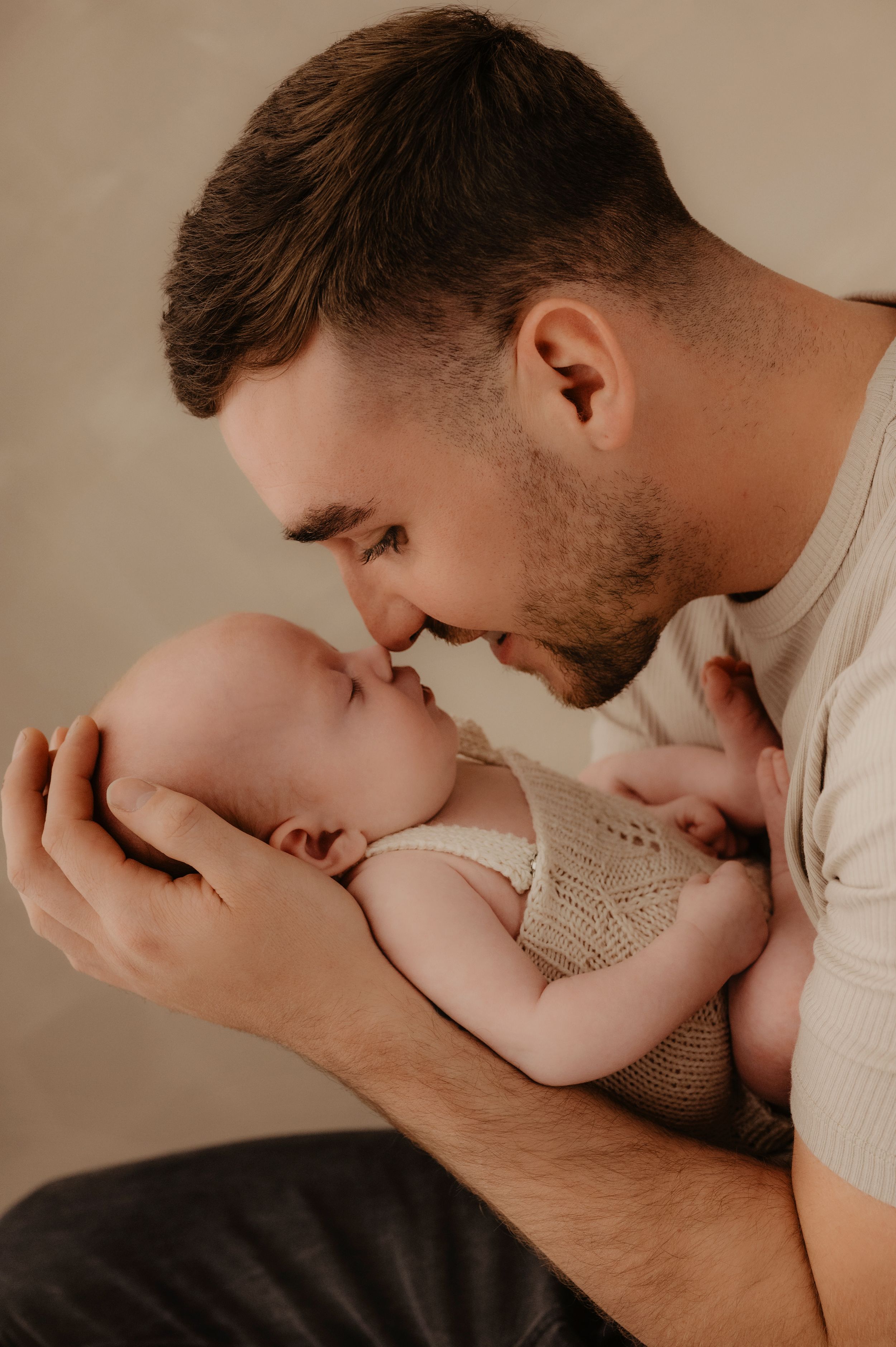 A tender moment between a father and newborn baby in a warm-toned portrait against a neutral background.