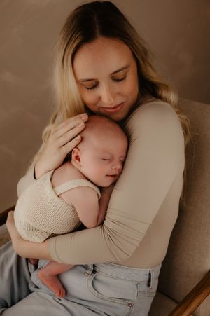 A tender moment shows a mother in jeans and beige top lovingly embracing her sleeping newborn on a brown chair.