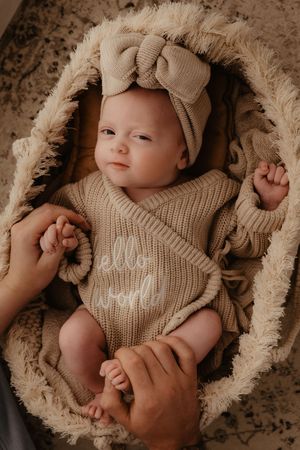 A baby wearing a beige knit romper and bow headband lies in a plush furry blanket while hands reach to hold them.