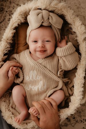Baby lying on cream furry blanket wearing beige knit romper and headband in professional studio photography layout.