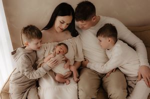 Family in neutral colored clothing cuddles together on beige couch while admiring newborn baby.