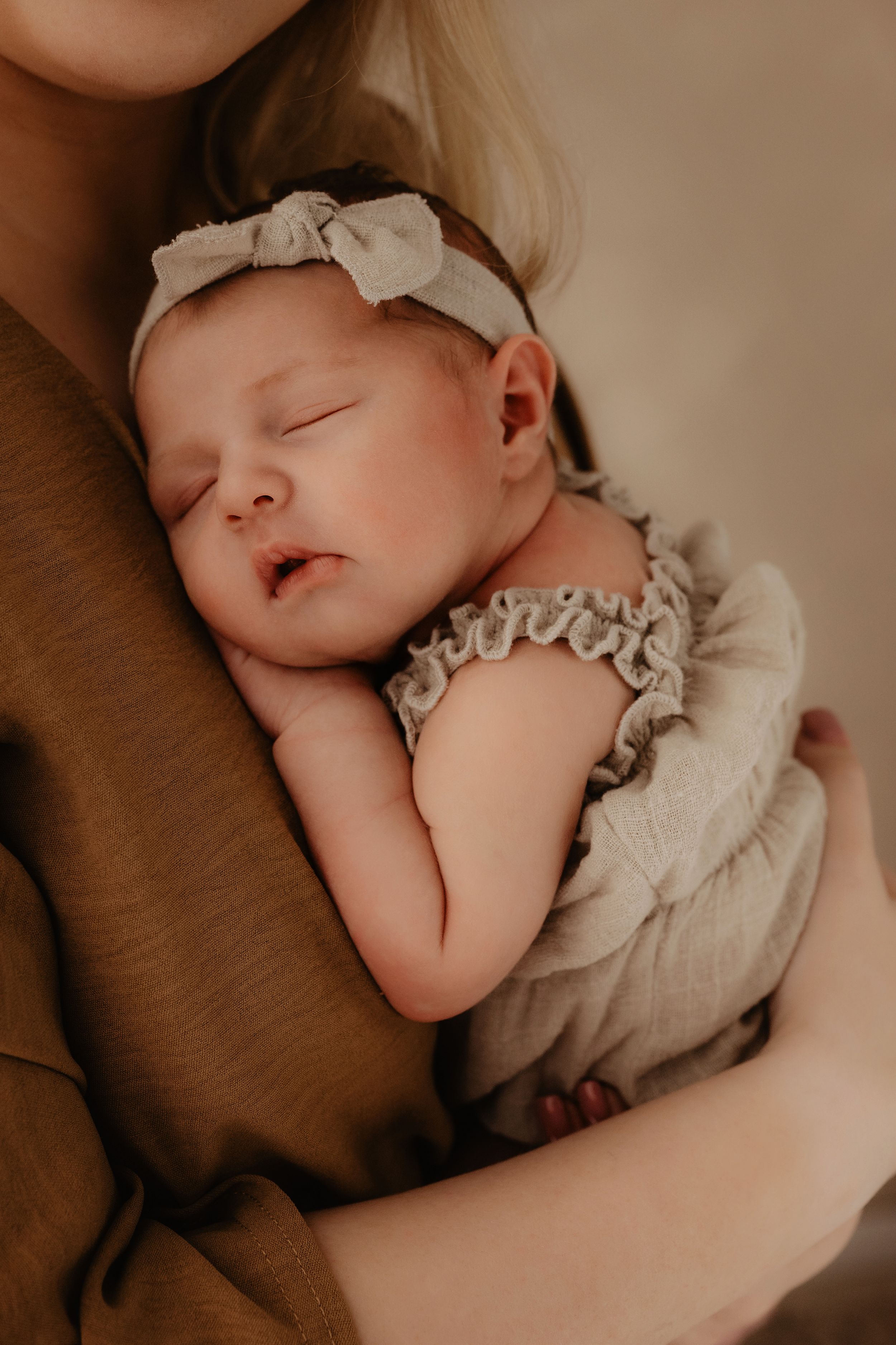 A peacefully sleeping baby wearing a light colored dress and bow headband nestles against someone in warm brown clothing.