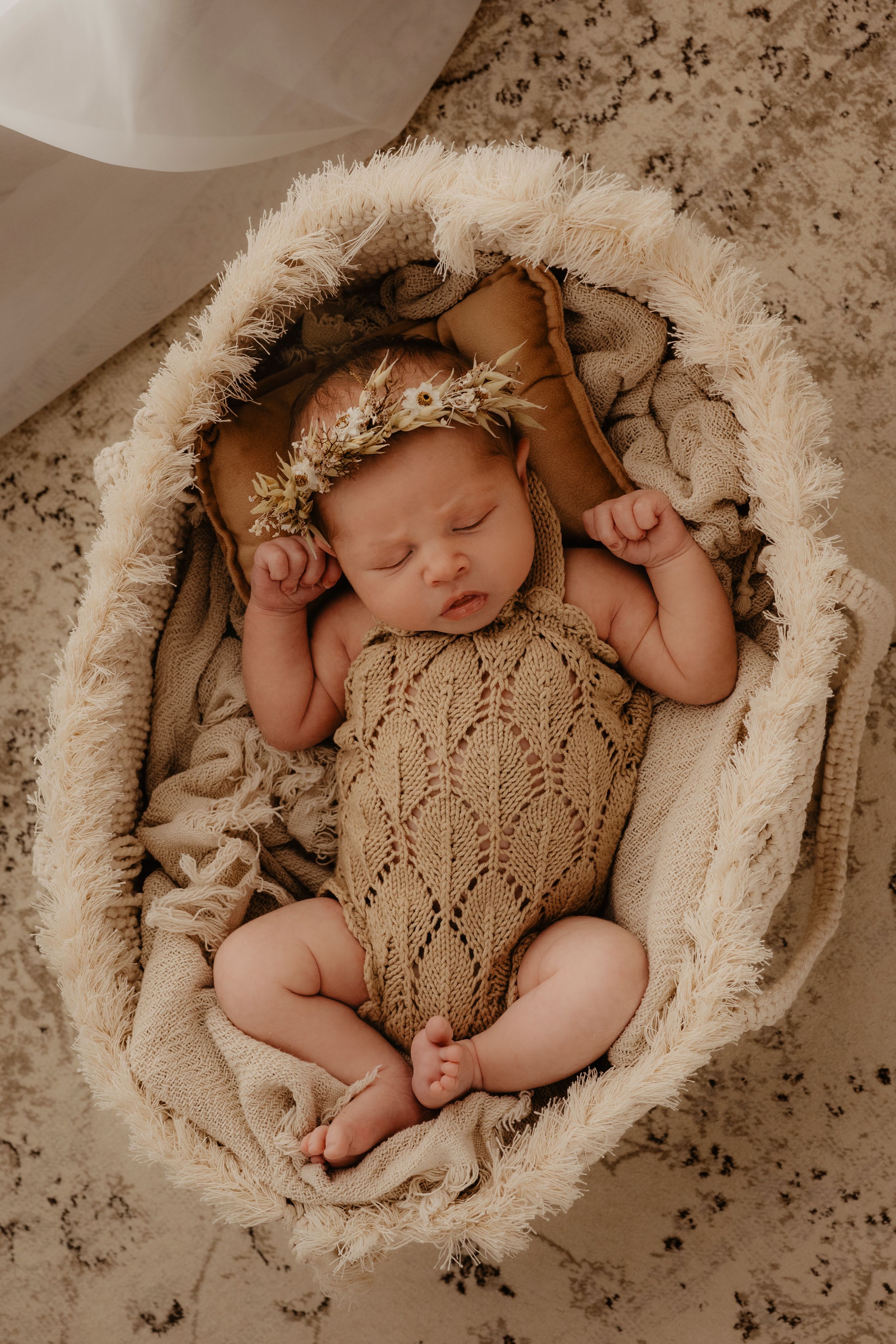 Sleeping newborn in a knitted beige romper with floral headband nestled in a furry cream-colored cocoon basket.