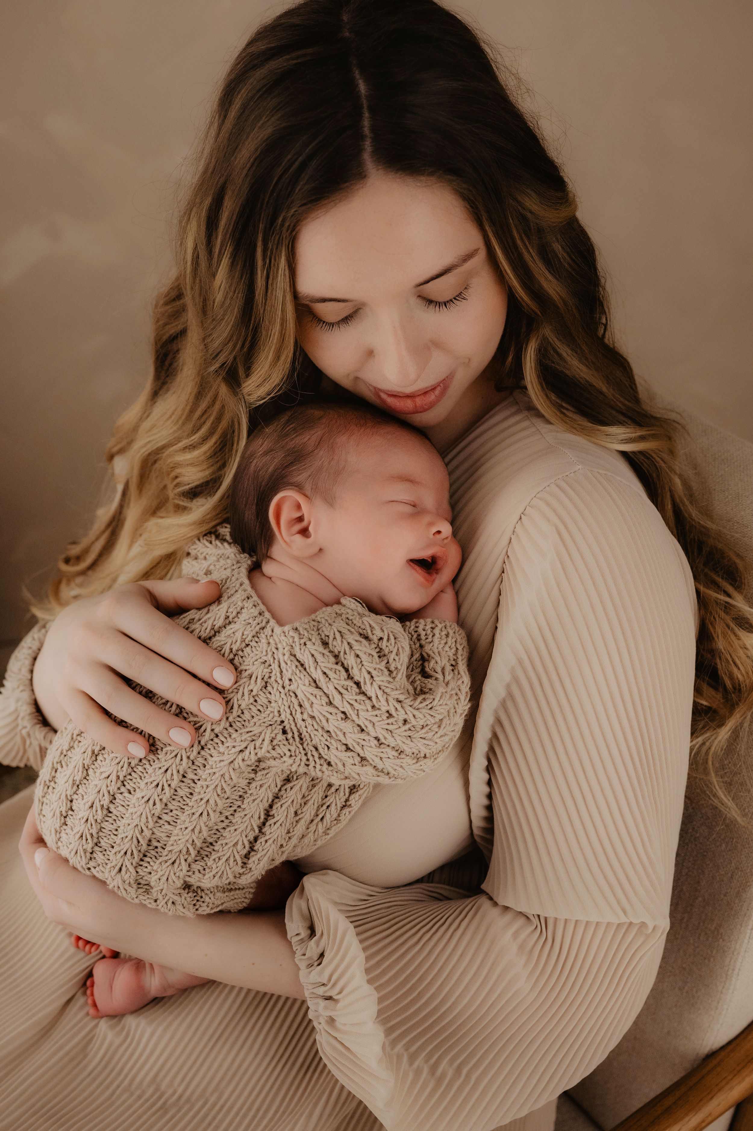 A tender moment between mother and newborn baby wearing knit sweaters in warm neutral tones during an intimate portrait session.