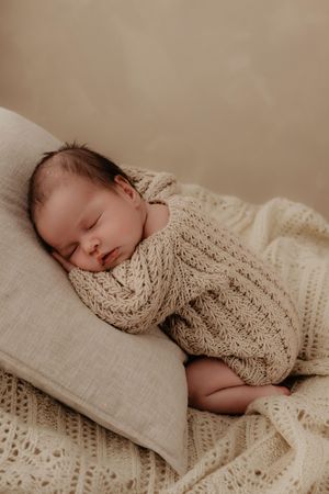A sleeping newborn wrapped in a cozy cream-colored knit blanket rests peacefully on soft beige bedding.