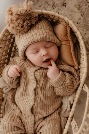 A sleeping baby wearing a cozy beige knit sweater and pom pom hat rests in a wicker basket.