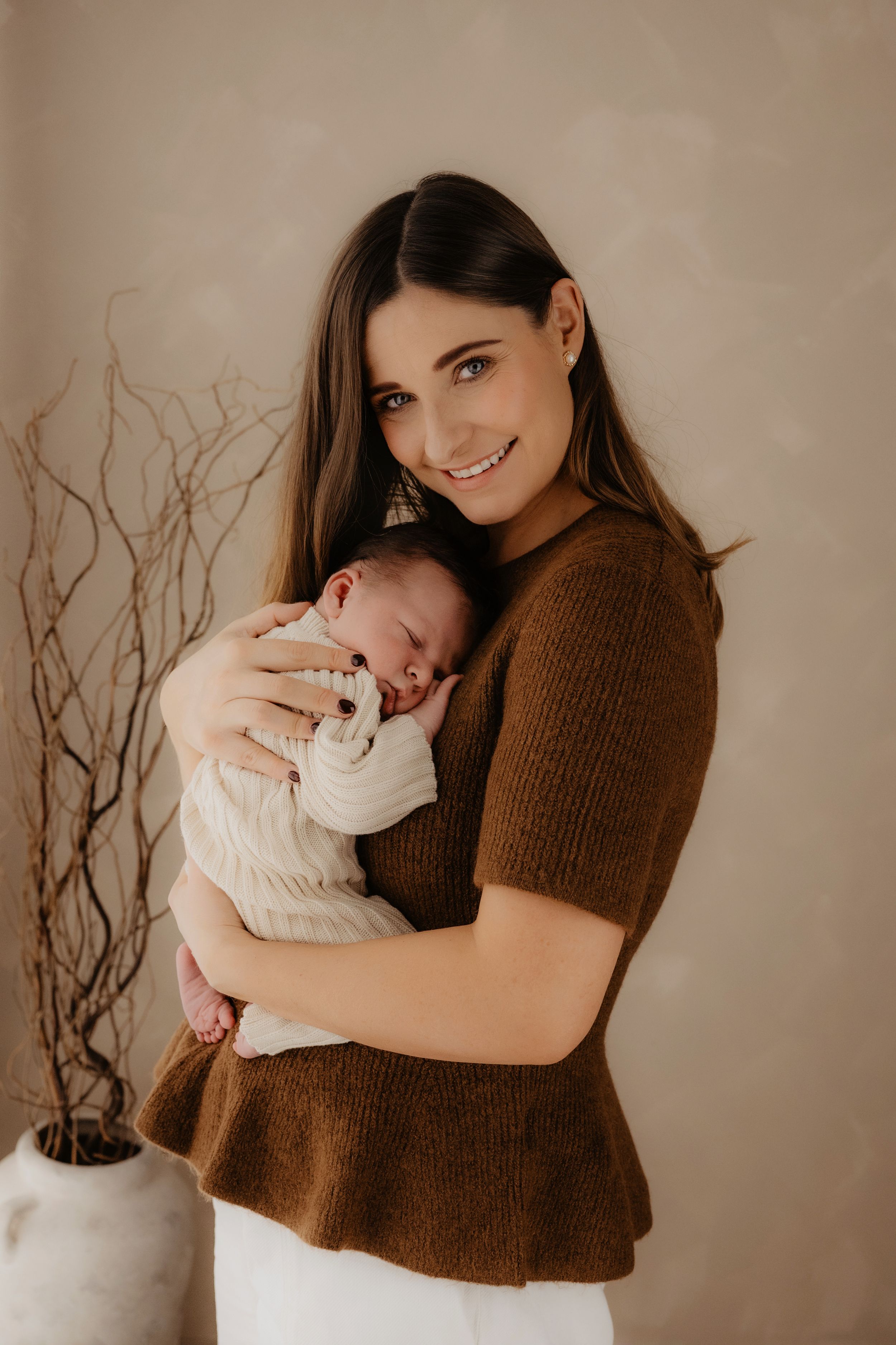 A person in a brown sweater holds a sleeping newborn baby against a neutral backdrop with decorative branches.