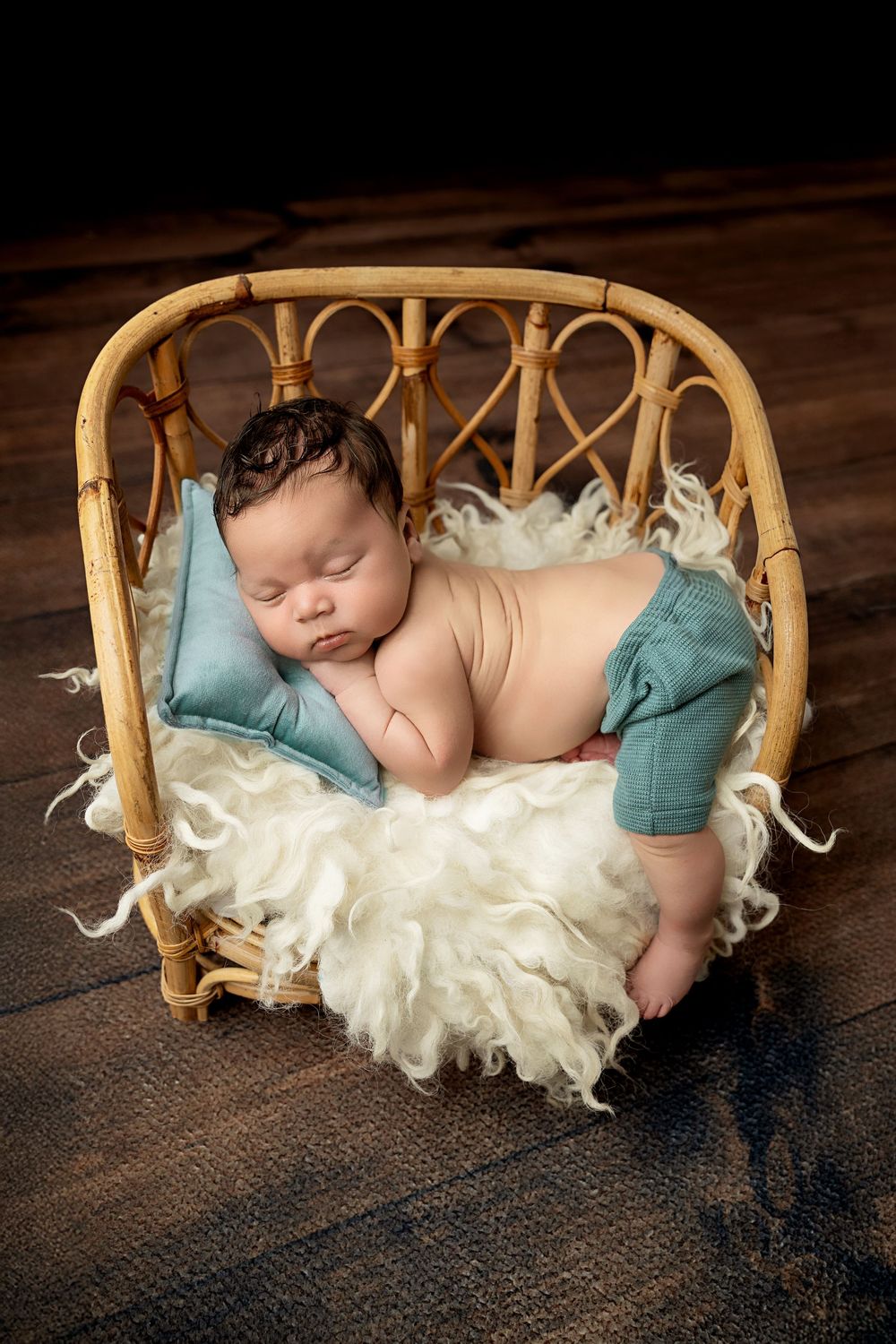 Sleeping newborn swaddled in cream fabric laying on a curved wooden bed prop with teal accents against dark backdrop.