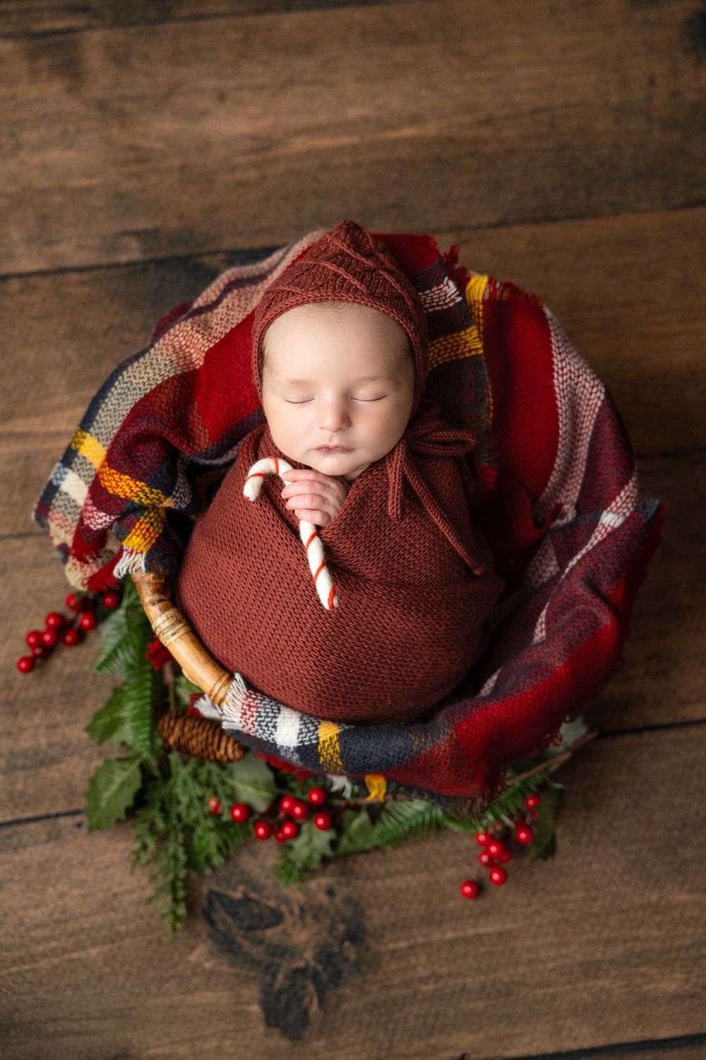 Sleeping newborn wrapped in brown knit swaddle with plaid blanket and red berries on wooden background.