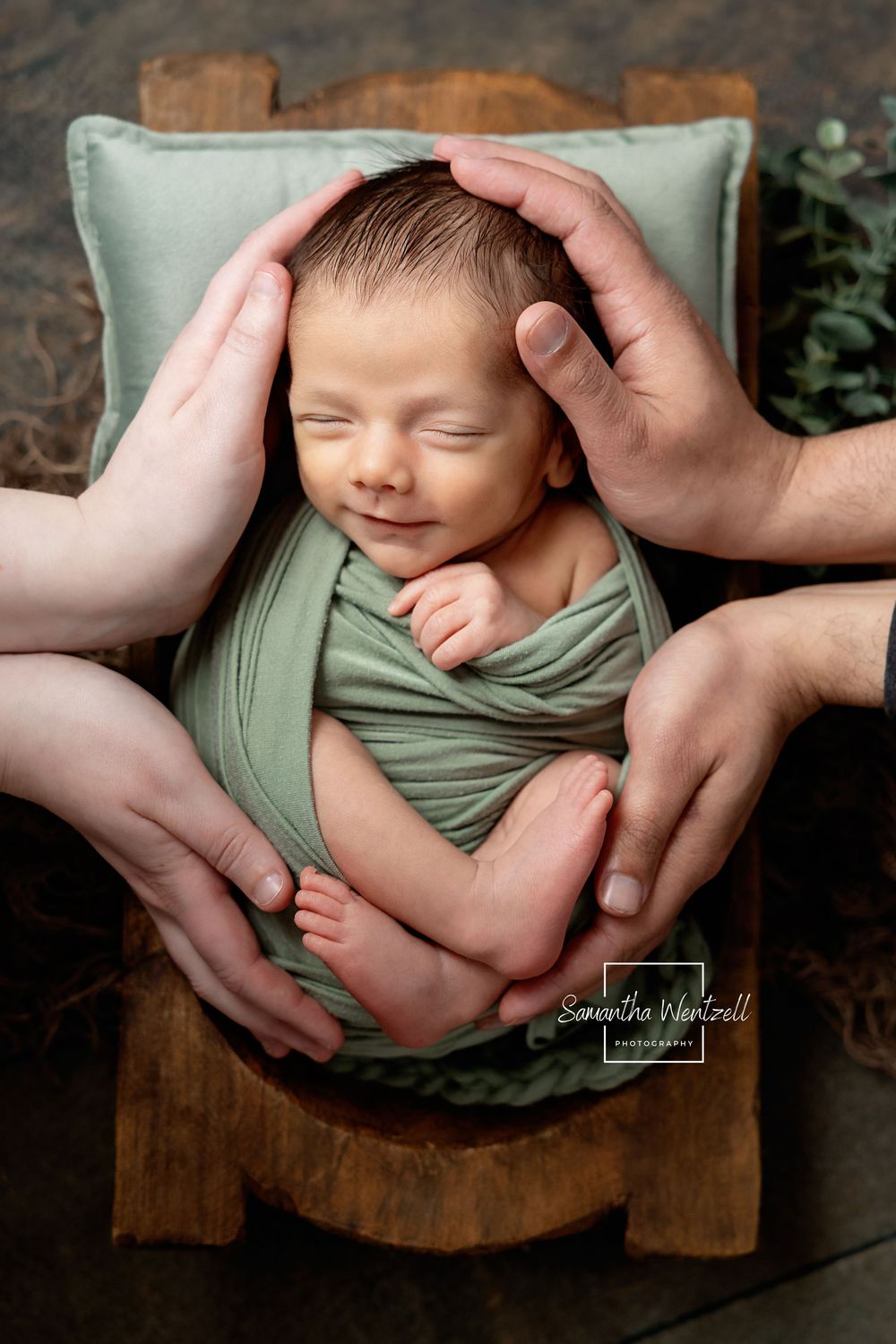 Newborn baby swaddled in green wrap sleeping in tiny wooden bed, cradled by parents hands in professional photo.