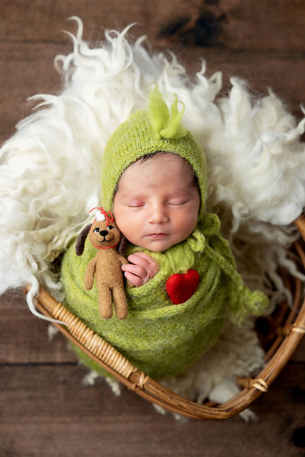 A sleeping baby swaddled in green knit wrap rests peacefully in a wicker basket with white fluffy decorative material.