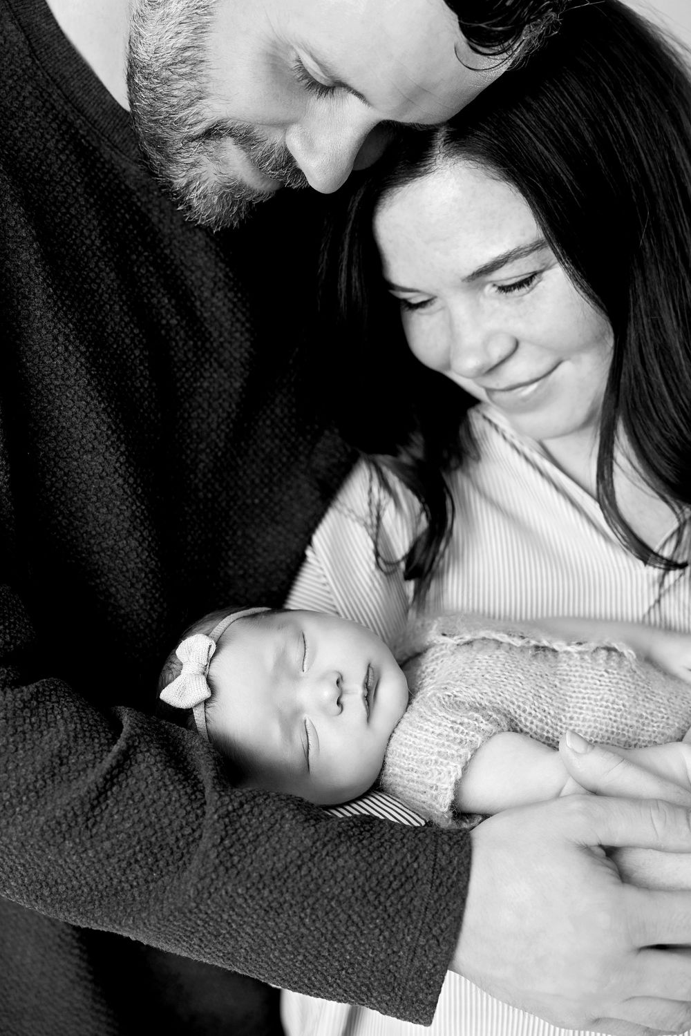 Black and white photo of parents lovingly gazing at their sleeping newborn baby girl with a bow headband.