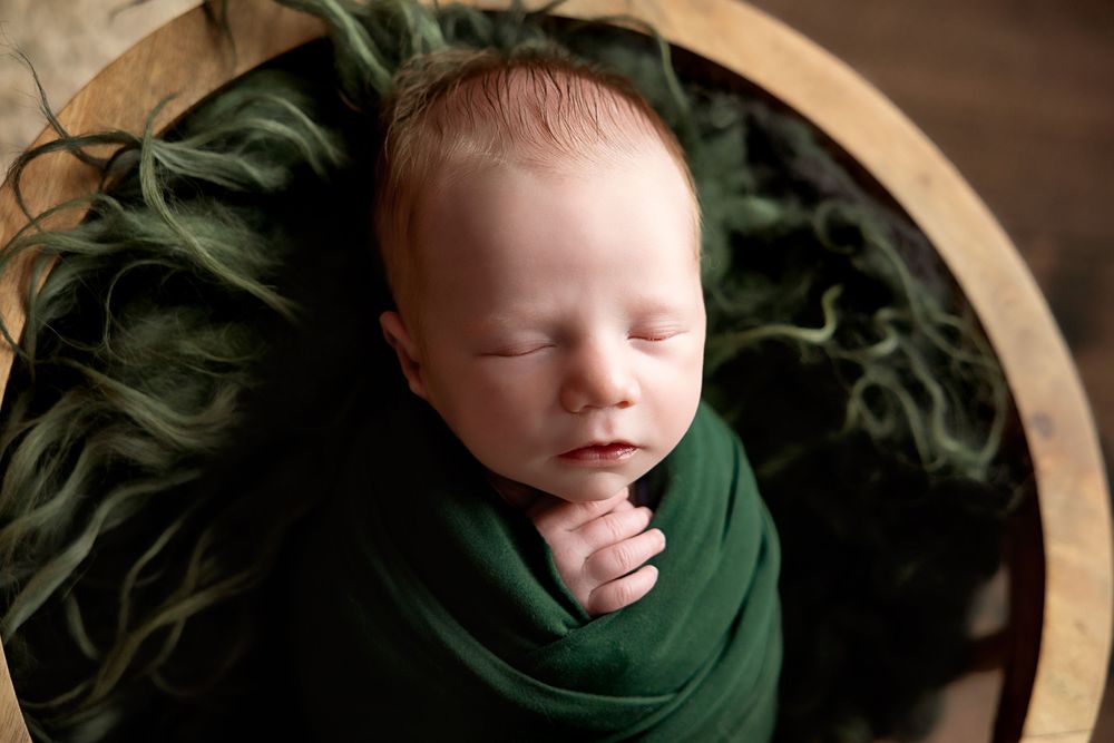 Newborn baby swaddled in a dark green wrap, sleeping peacefully in a round wooden bowl with green floral accents.