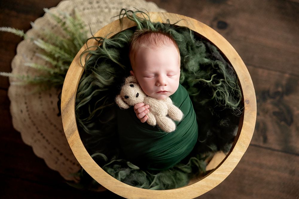 Newborn baby wrapped in green swaddle sleeping in wooden bowl with teddy bear and fern leaves.