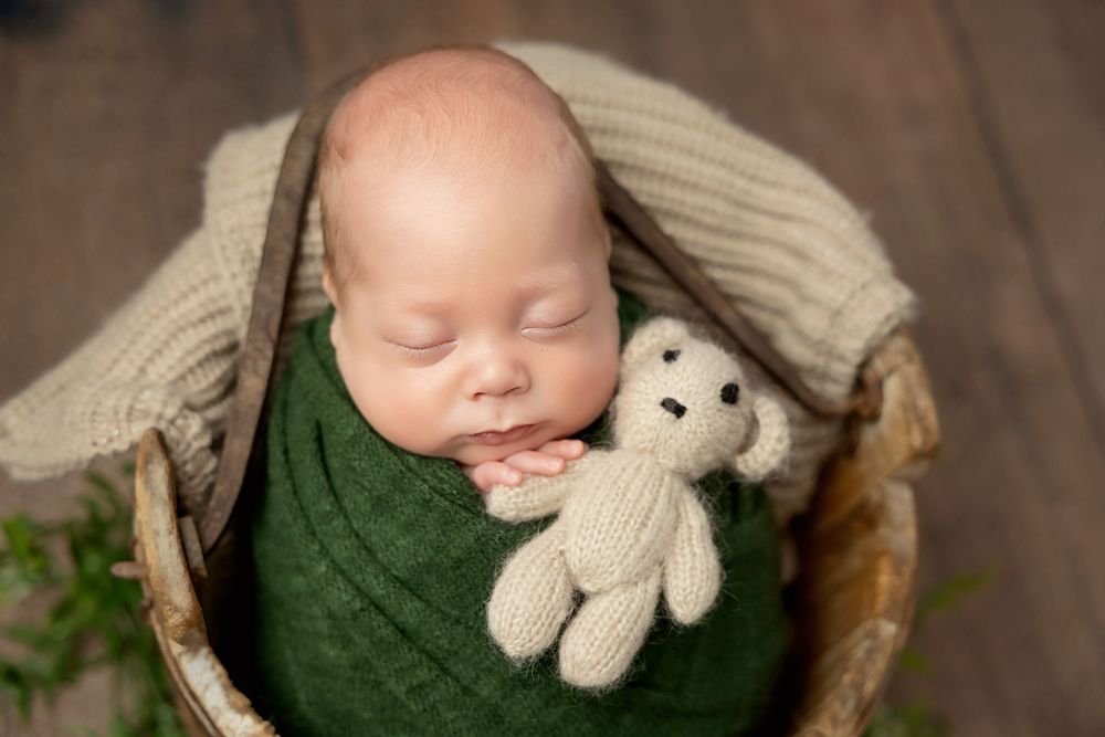 A sleeping baby in a rustic basket wears a green knit wrap while cuddling a plush elephant toy against a wooden backdrop.
