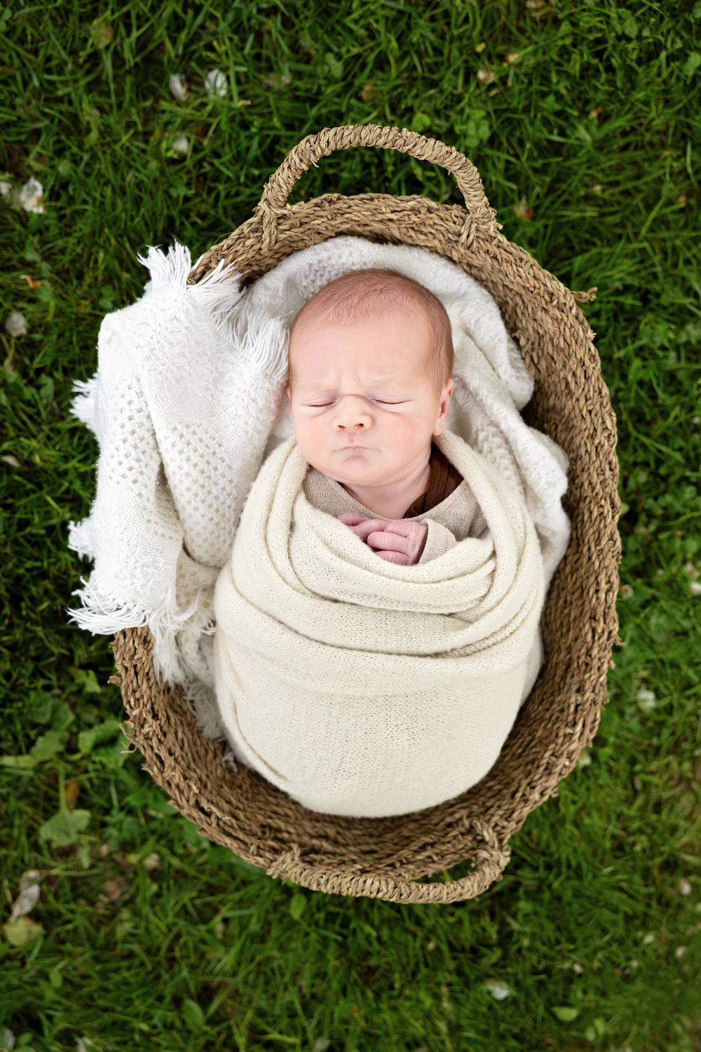 Newborn baby swaddled in cream wrap sleeping peacefully in a wicker basket on green grass outdoors.