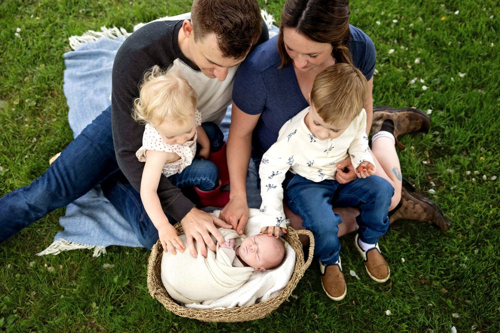 Family of four sitting on grass, parents admiring newborn baby in basket with toddler siblings outdoors.
