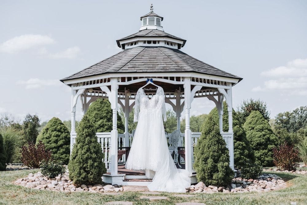 Wedding dress hanging in a white gazebo surrounded by evergreen trees on a sunny day.