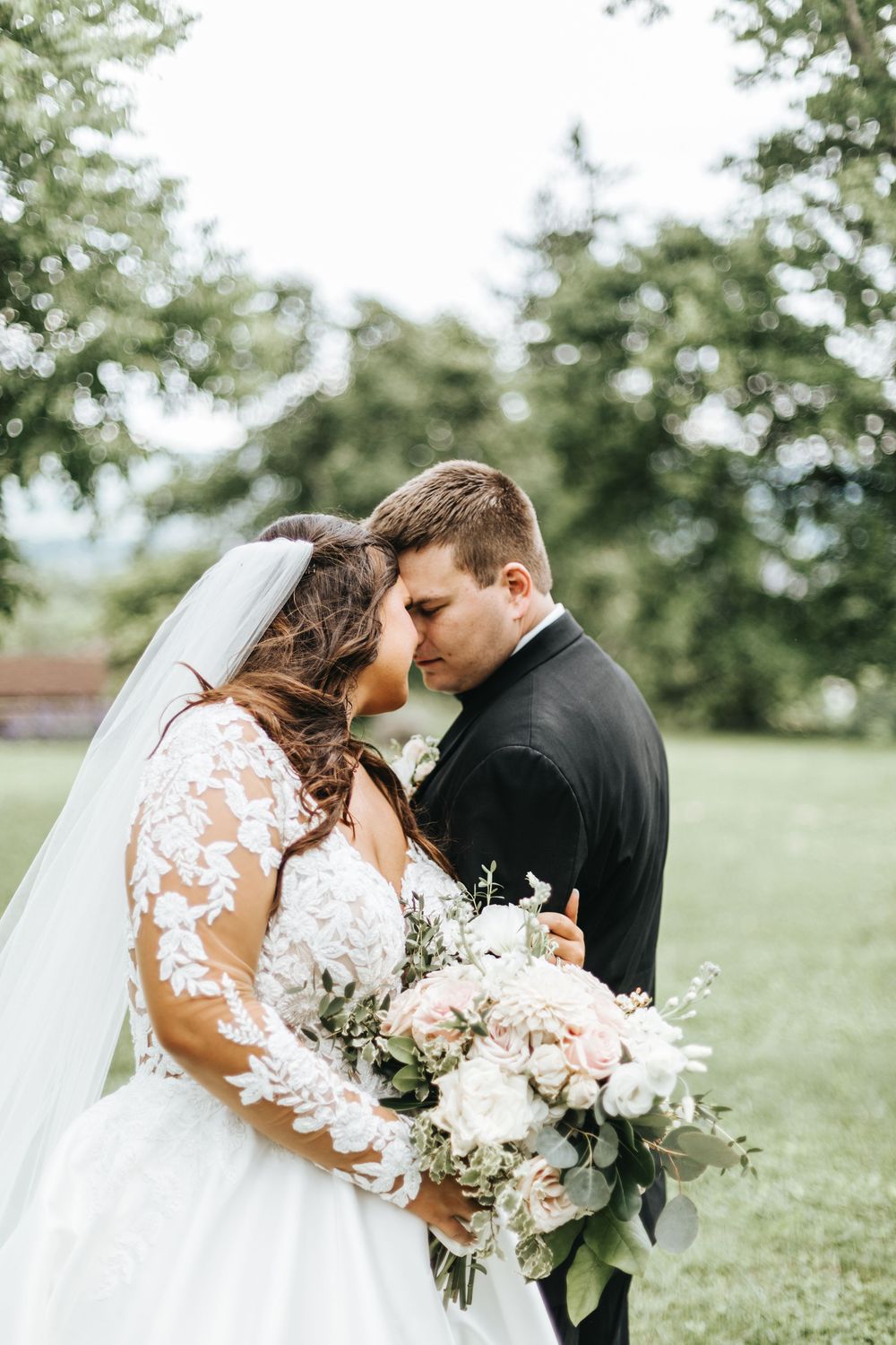 Bride and groom share romantic forehead touch outdoors, bride holding elegant white and blush bouquet in lace gown.