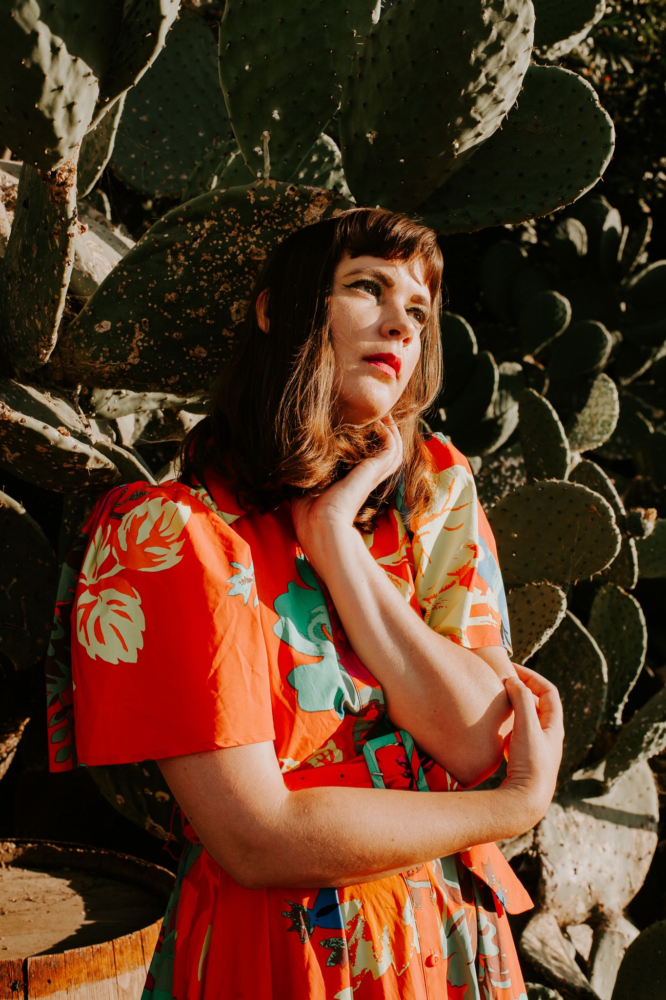 A woman looking away wearing an orange dress with flowers with a cactus in the background.