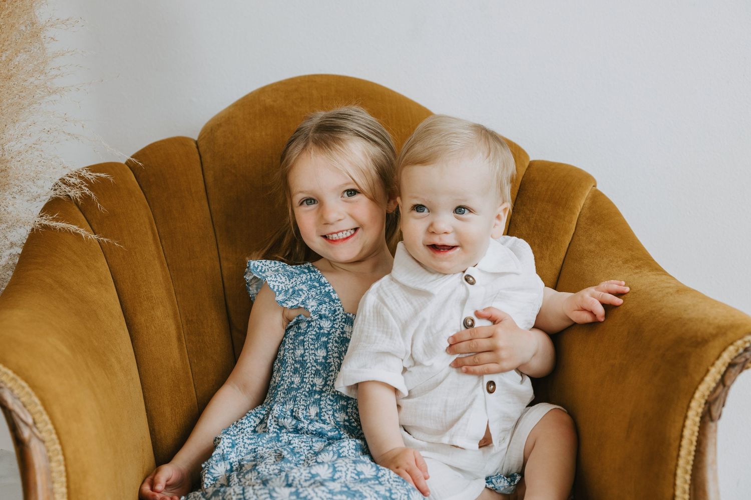 Two children sit together on a vintage gold velvet armchair during a photo session.