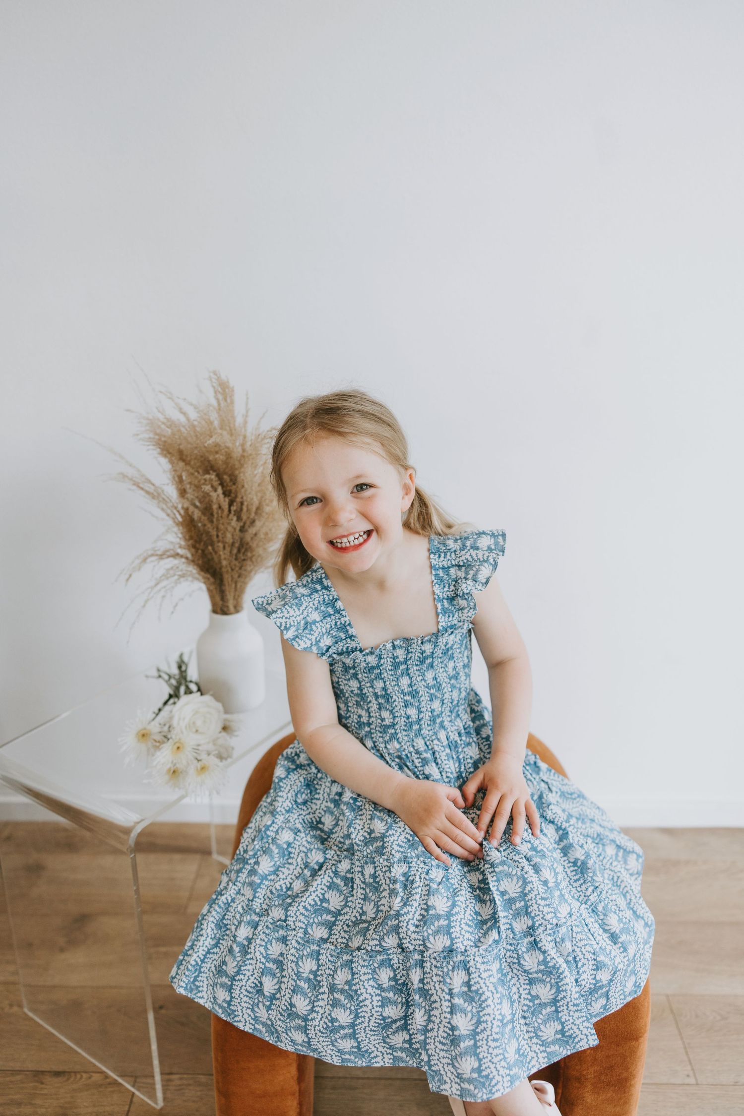 Child in blue floral dress sits on wooden chair next to dried pampas grass arrangement.