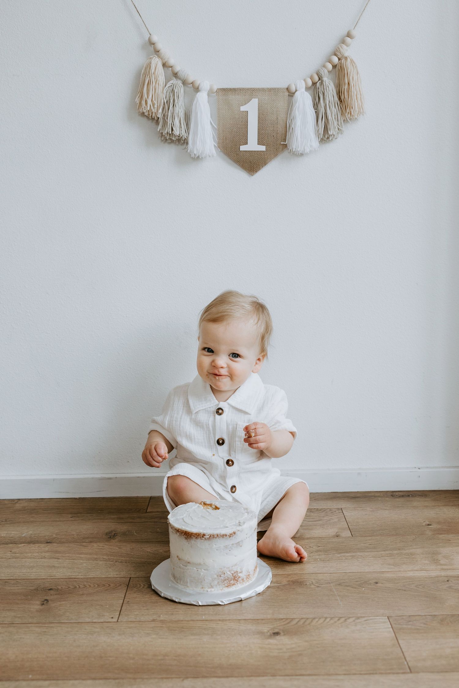 Baby sits under number one banner with small white cake on wooden floor.