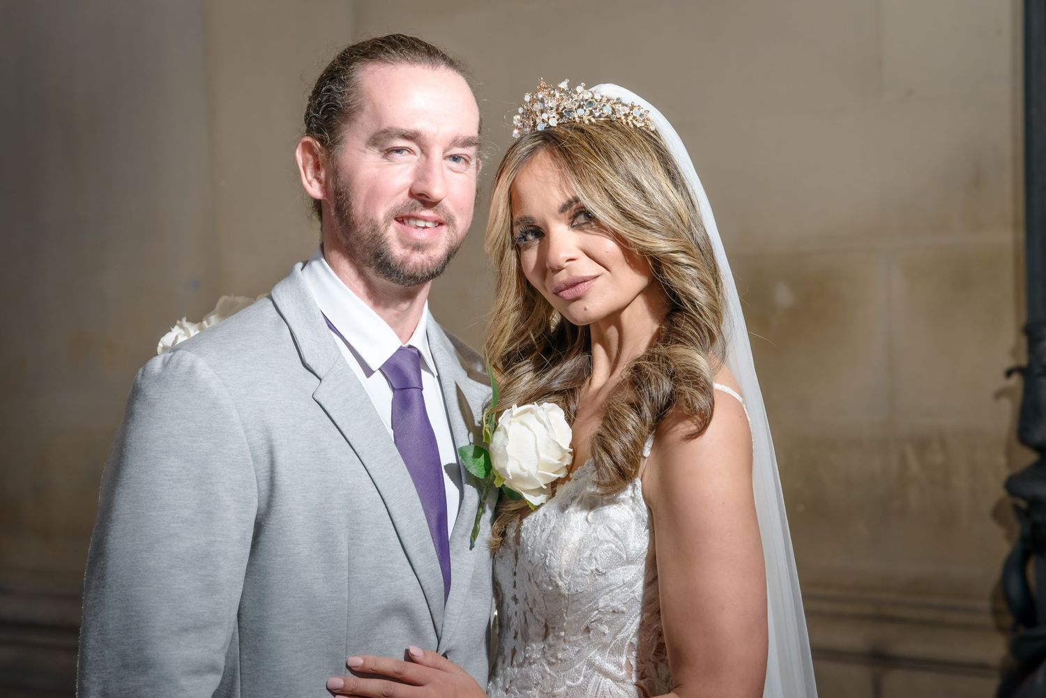 Couple in wedding attire stand together, with bride wearing lace dress and crystal tiara, groom in light gray suit and purple tie.
