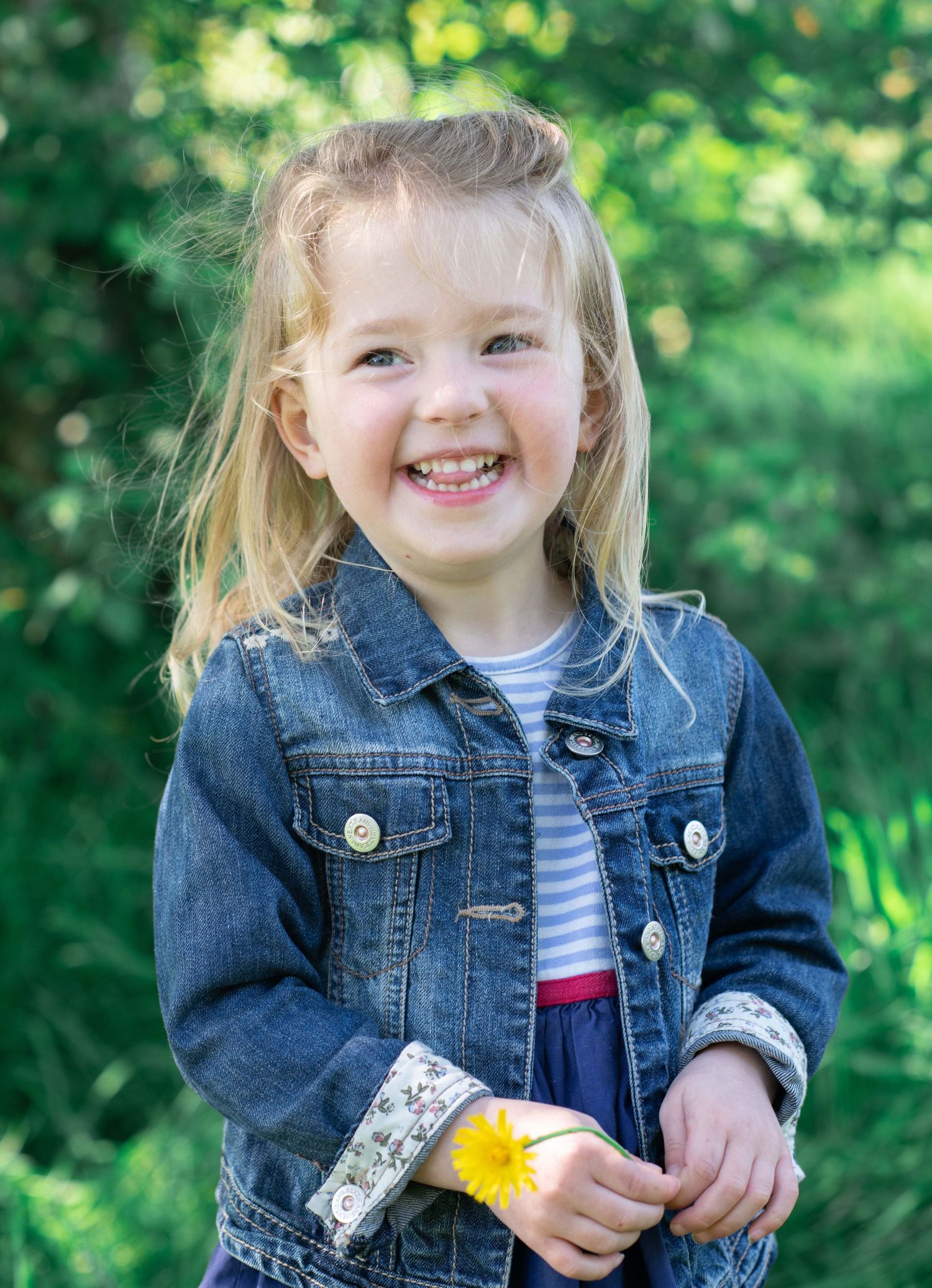A cheerful young child in denim jacket stands outdoors holding a yellow dandelion flower against green foliage background.
