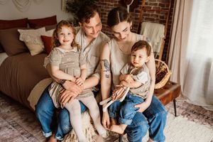 A young family of four sits together in a cozy bedroom, parents holding two small children in casual earth-tone outfits.