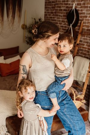 A young mother with tattoos holds her toddler son while her little daughter stands beside her in a cozy room.