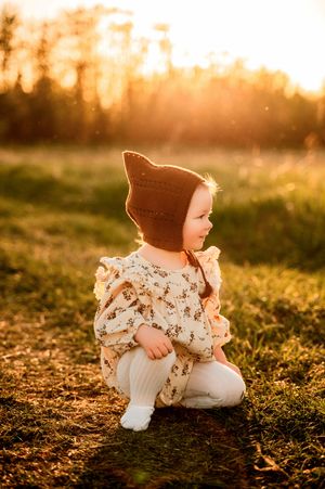 A small figure wearing a knit hat and floral dress crouches in a field at golden hour as sunlight filters through trees.