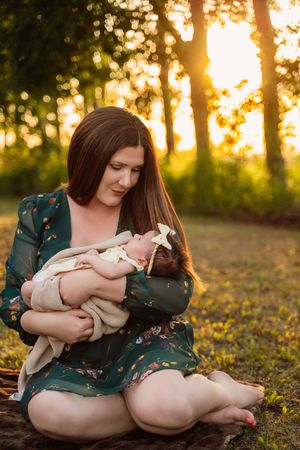 Woman in floral dress cuddles newborn baby on grassy hill during sunset in the woods.