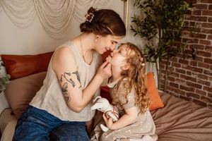 A tattooed mother tenderly touches noses with her curly-haired toddler daughter while sitting on a bed.