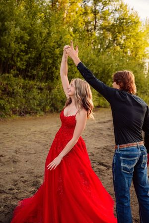 Two people in casual attire dance joyfully together against an autumn backdrop at sunset, one wearing a flowing red dress.