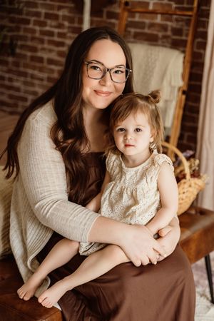 A woman with glasses holds a toddler girl on her lap, both wearing neutral tones, seated in a warmly lit brick-walled studio.