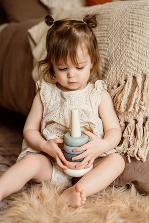 Toddler girl in white knit dress playing with stacking toy rings on cozy fur rug with cushions.