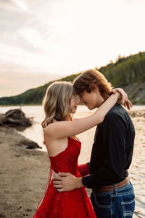 Two figures embrace in a romantic moment on a beach at sunset, with one wearing a red dress against a coastal backdrop.