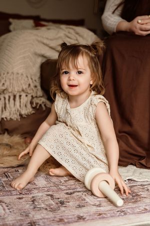 Smiling toddler girl in cream dress sitting on rug holding wooden toy with brown bedding in background.