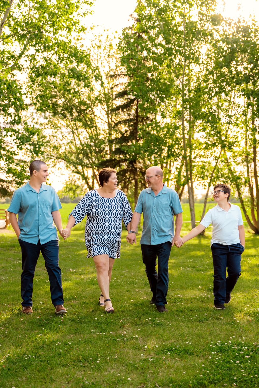 Family of four holding hands and walking together in a sunny green park surrounded by lush trees.