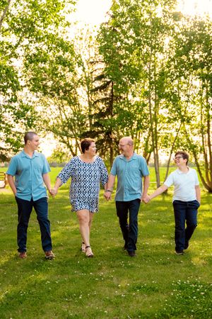 Family of four holding hands and walking together in a sunny green park surrounded by lush trees.