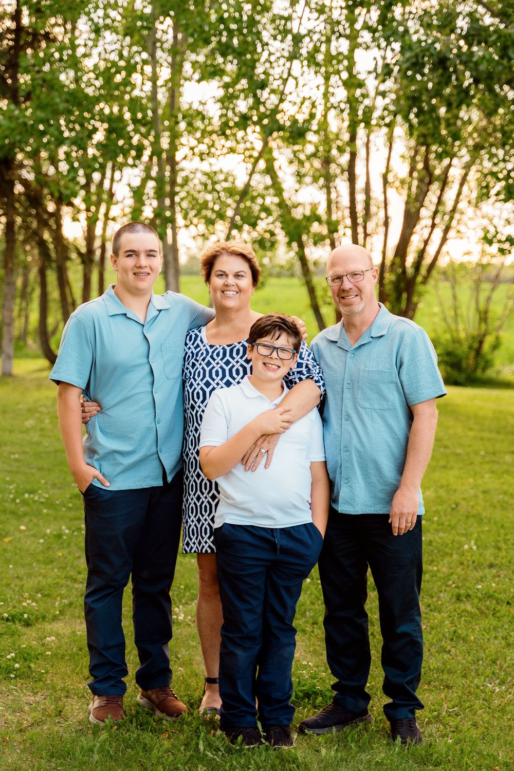 Family of four posing outdoors on green grass with trees, wearing coordinating blue outfits for a portrait session.