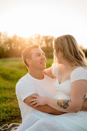 A couple in white outfits embraces while sitting in a grassy field during golden hour sunset.