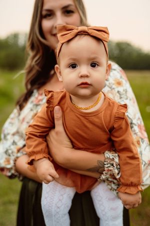 Four photos show a baby wearing a brown outfit with a matching bow headband being held up outdoors at sunset.