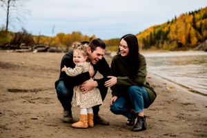 A family crouches together on a sandy beach in autumn with golden trees lining the shore in the background.
