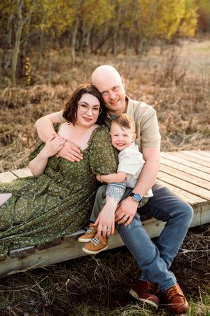A happy family of three poses outdoors on a wooden platform, mom in green floral dress, dad, and young son smiling together.