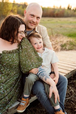 A happy family of three sits on a wooden dock outdoors, with a bald dad, mom in green floral dress, and smiling toddler son.