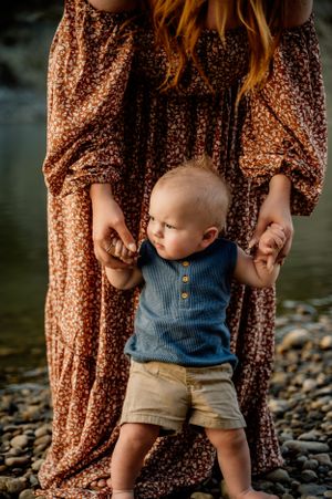 A baby wearing a blue tank top and khaki shorts stands on a rocky beach while holding hands with someone in a floral dress.